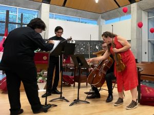 Woman in orange dress points to sheet music for student cellist while holding violin