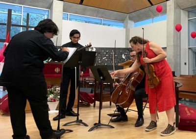 Woman in orange dress points to sheet music for student cellist while holding violin