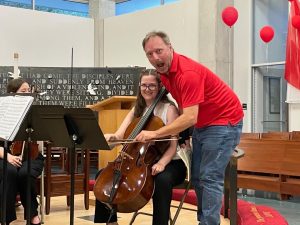 Man shows mock surprise while holding smiling student's bow on student's cello