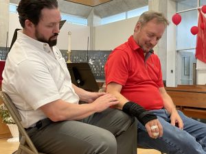 Bearded man in white shirt touches the elbow of another man in wrist brace and bandages in a diagnostic manner