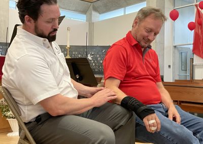 Bearded man in white shirt touches the elbow of another man in wrist brace and bandages in a diagnostic manner
