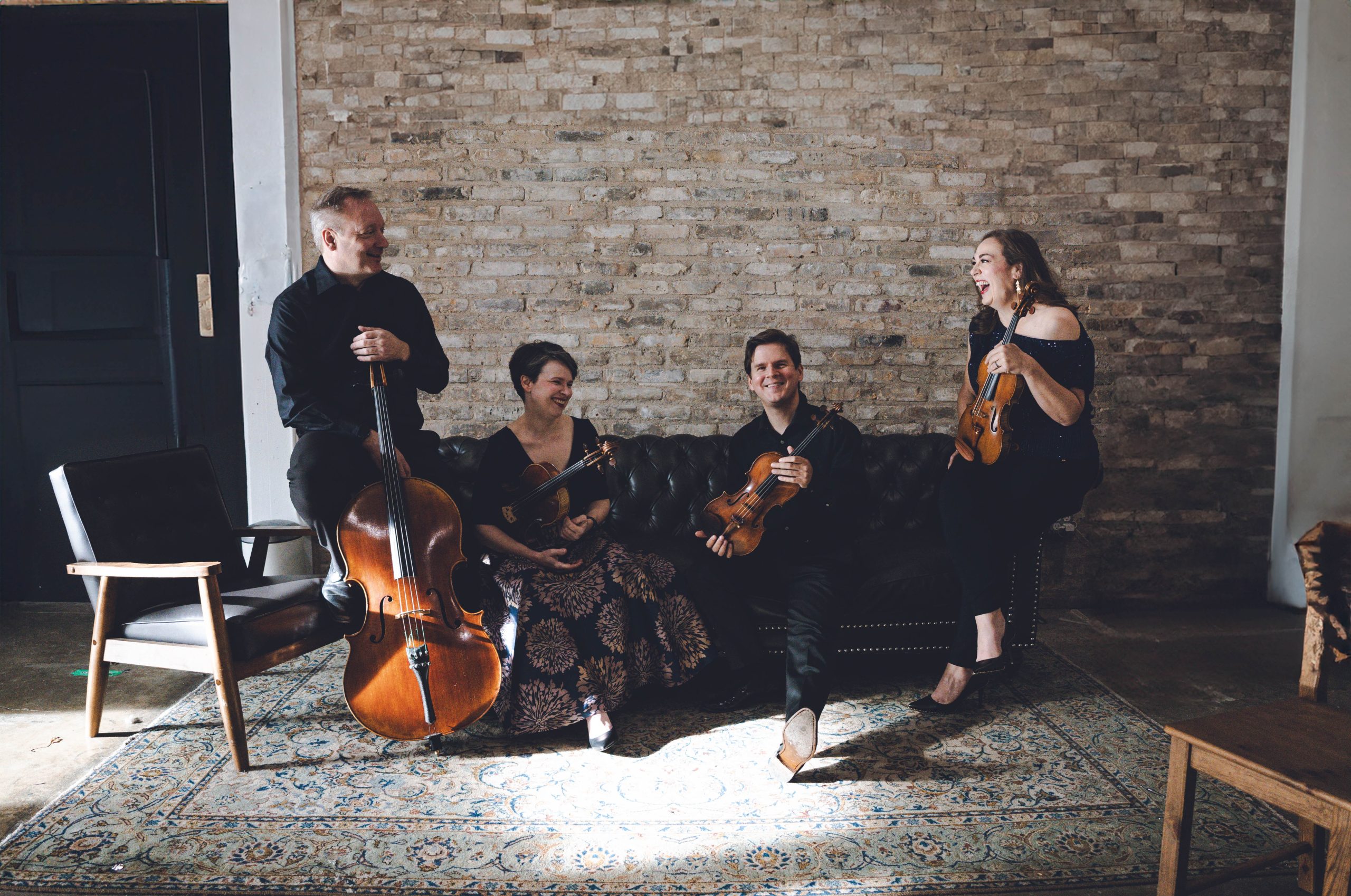 Far left, Man with cello perches on the arm of leather couch, looking at colleagues to the right. Woman in long dress seated on the couch holds viola, faces to the right, is laughing. Man to right is holding violin, also seated on the couch. Woman holding violin is perched on the right arm of the leather couch, laughing heartily. All before a brick wall