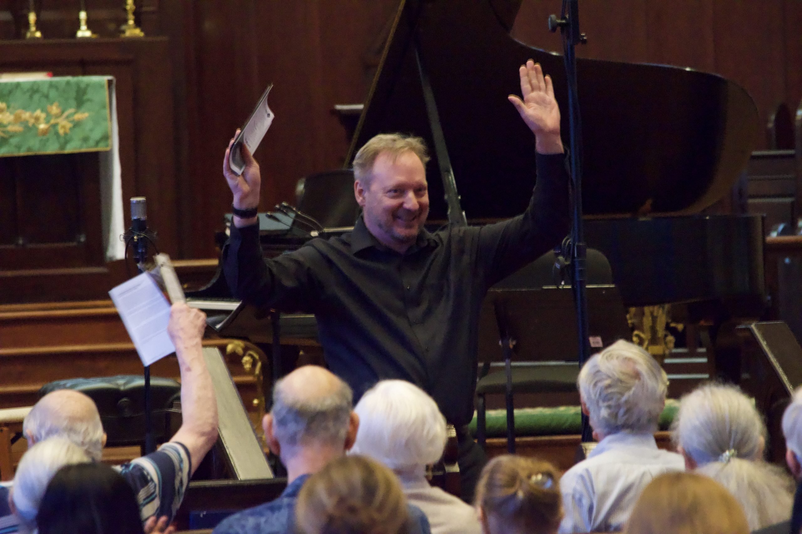 Man in a black shirt holds hands up smiling and holding a program interacting with an audience