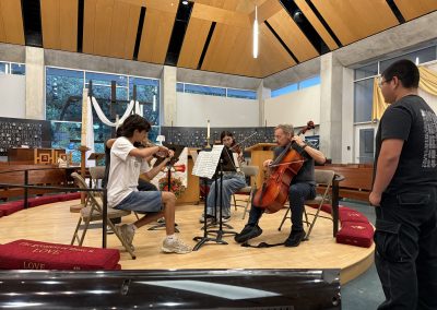Teacher instructs from within the young string quartet, playing cello while the young cellist observes standing